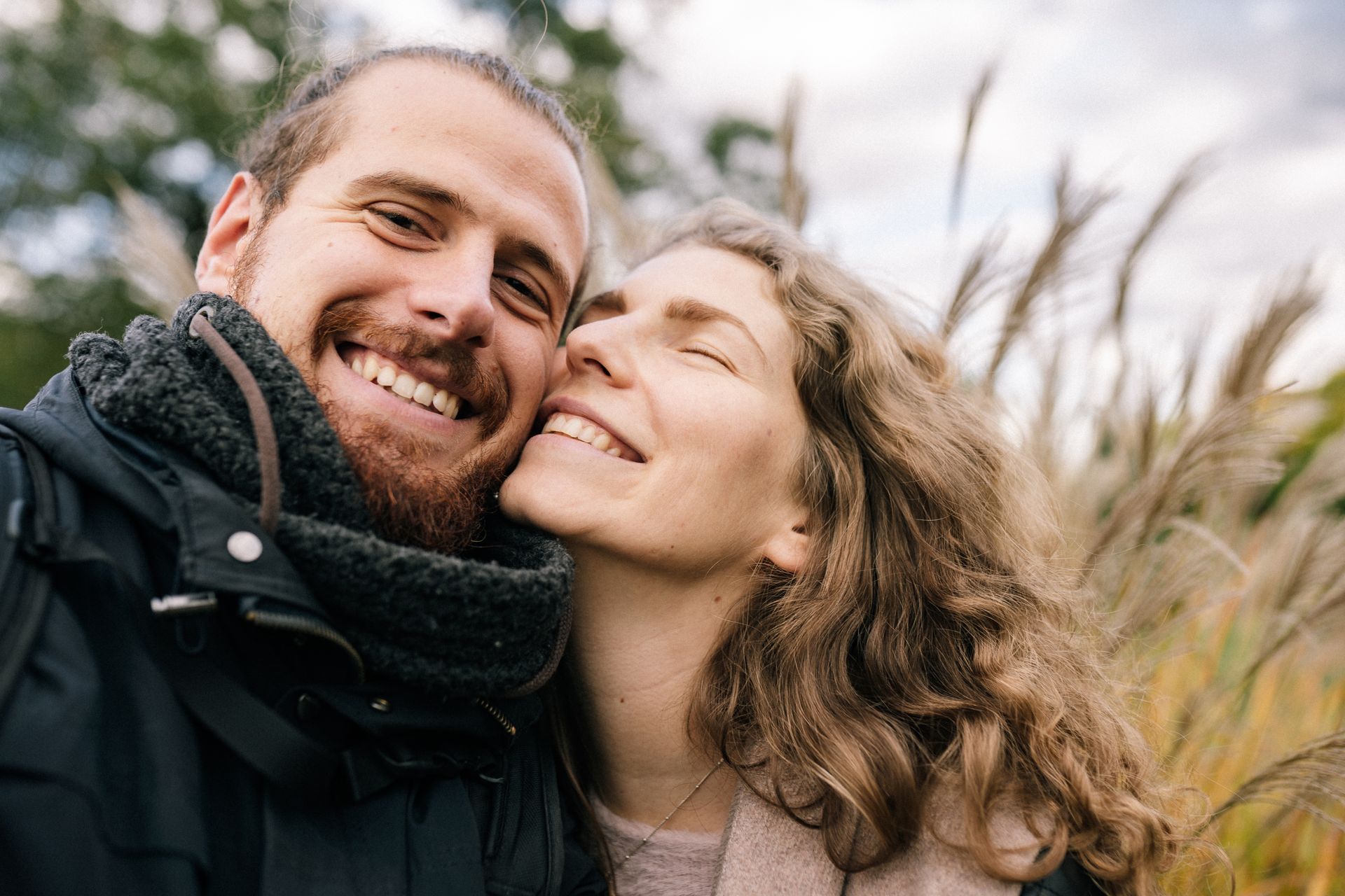 A smiling couple taking a close-up selfie outdoors in a wooded, wintery setting.